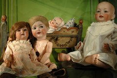 Puppen: vorne zwei Kinder, daneben ein Baby, im Hintergrund weiteres Baby im Bettchen