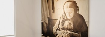 Eine Ausstellungsansicht mit einer Vitrine mit Objekten. Im Hintergrund ist ein Foto einer älteren Frau zu sehen.