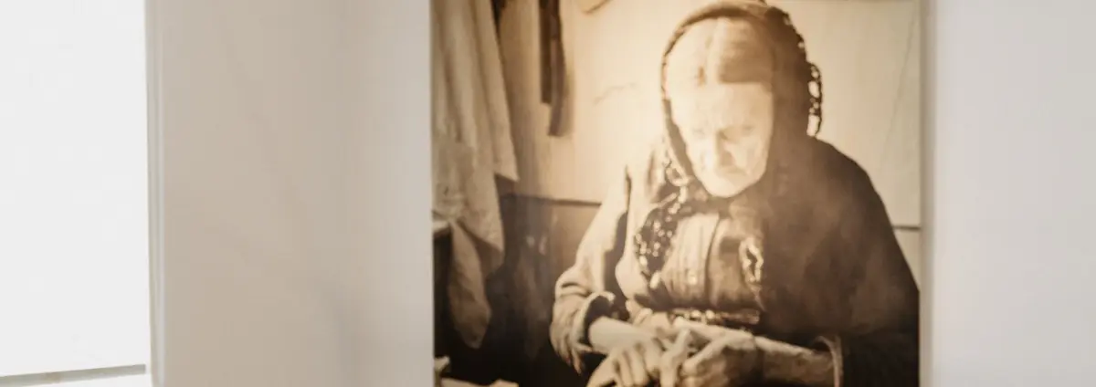 Eine Ausstellungsansicht mit einer Vitrine mit Objekten. Im Hintergrund ist ein Foto einer älteren Frau zu sehen.