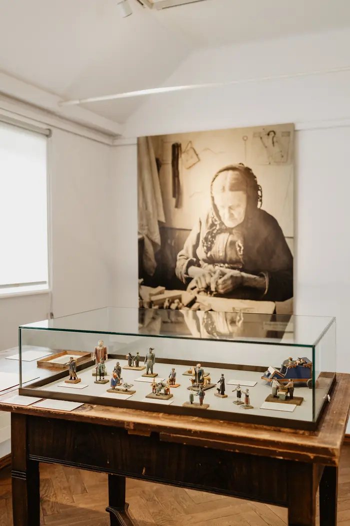 Eine Ausstellungsansicht mit einer Vitrine mit Objekten. Im Hintergrund ist ein Foto einer älteren Frau zu sehen.