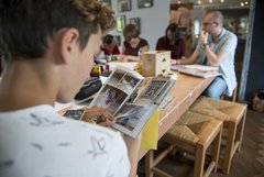 Junge blättert durch ein Heft mit Graffiti, mit am Tisch sitzen bastelnde Personen