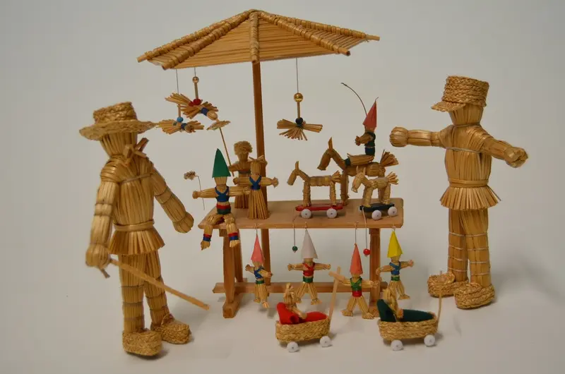 Zwei Strohfiguren und ein Tisch mit Schirm, auf und unter dem kleine stroh-Objekte stehen.