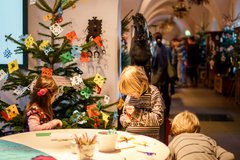 Kinder basteln an einem Tisch, dahinter ein Weihnachtsbaum mit Papierschmuck