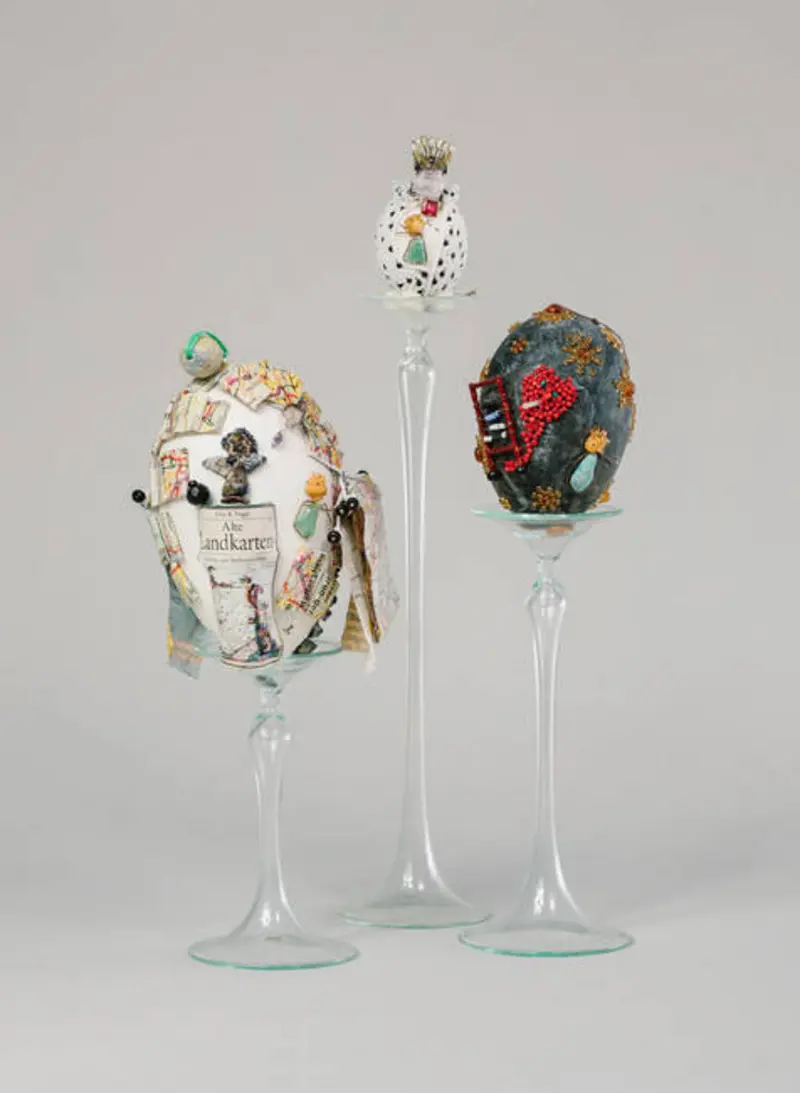 drei reich verzierte, Personen darstellende Eier auf Glasständern